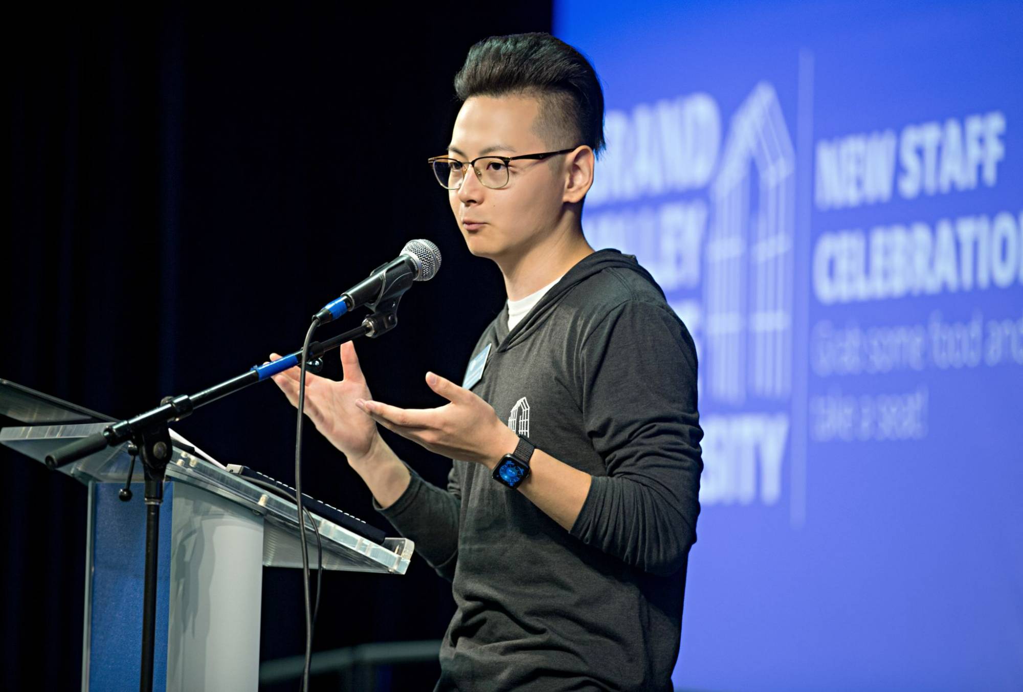 A young man talking into a microphone on stage. There is a podium in front of him.